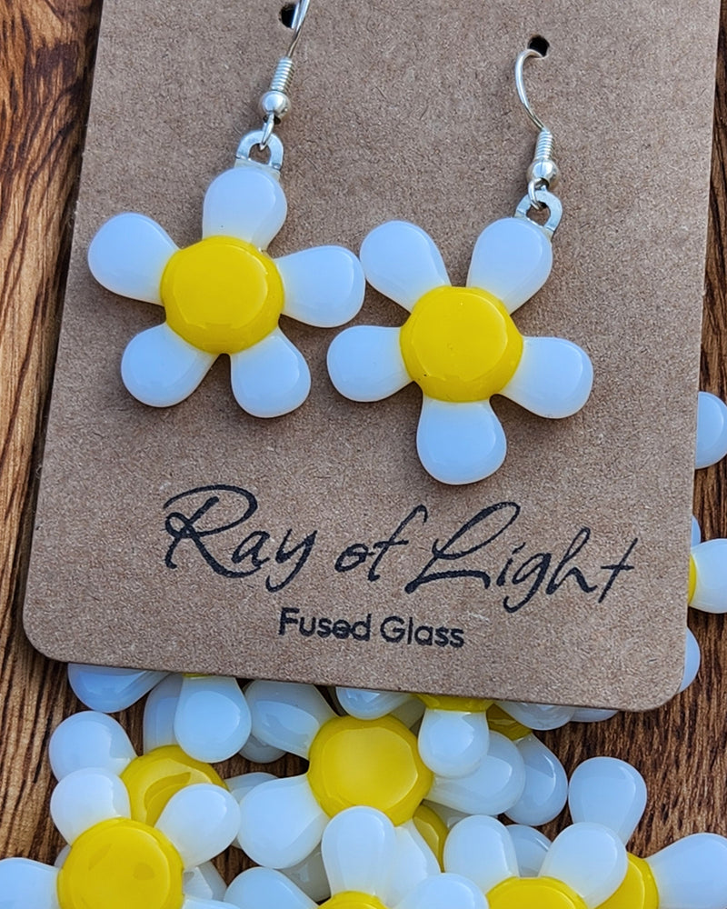 White and yellow glass daisy earrings hanging from a cardboard product display with Ray of Light Fused Glass printed at the bottom.