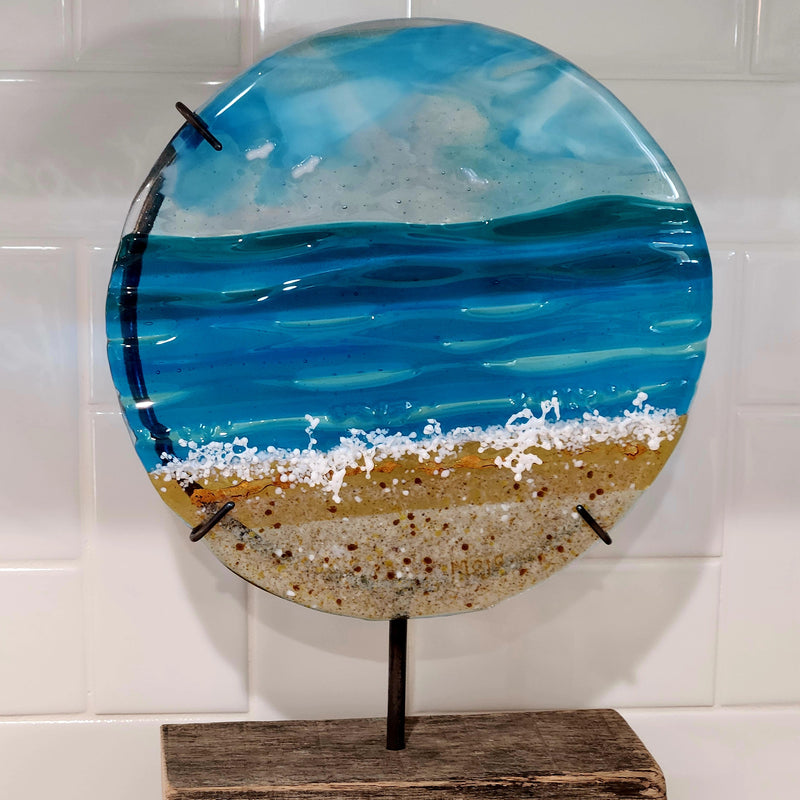 Memorial glass art panel in metal frame on wood stand. Glass features the ocean and the beach with cremation remains inside.