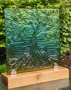 A square glass art tile featuring a raised, textured tree design in iridescent green, blue, and purple tones, displayed on a light wood stand outdoors with green foliage in the background.