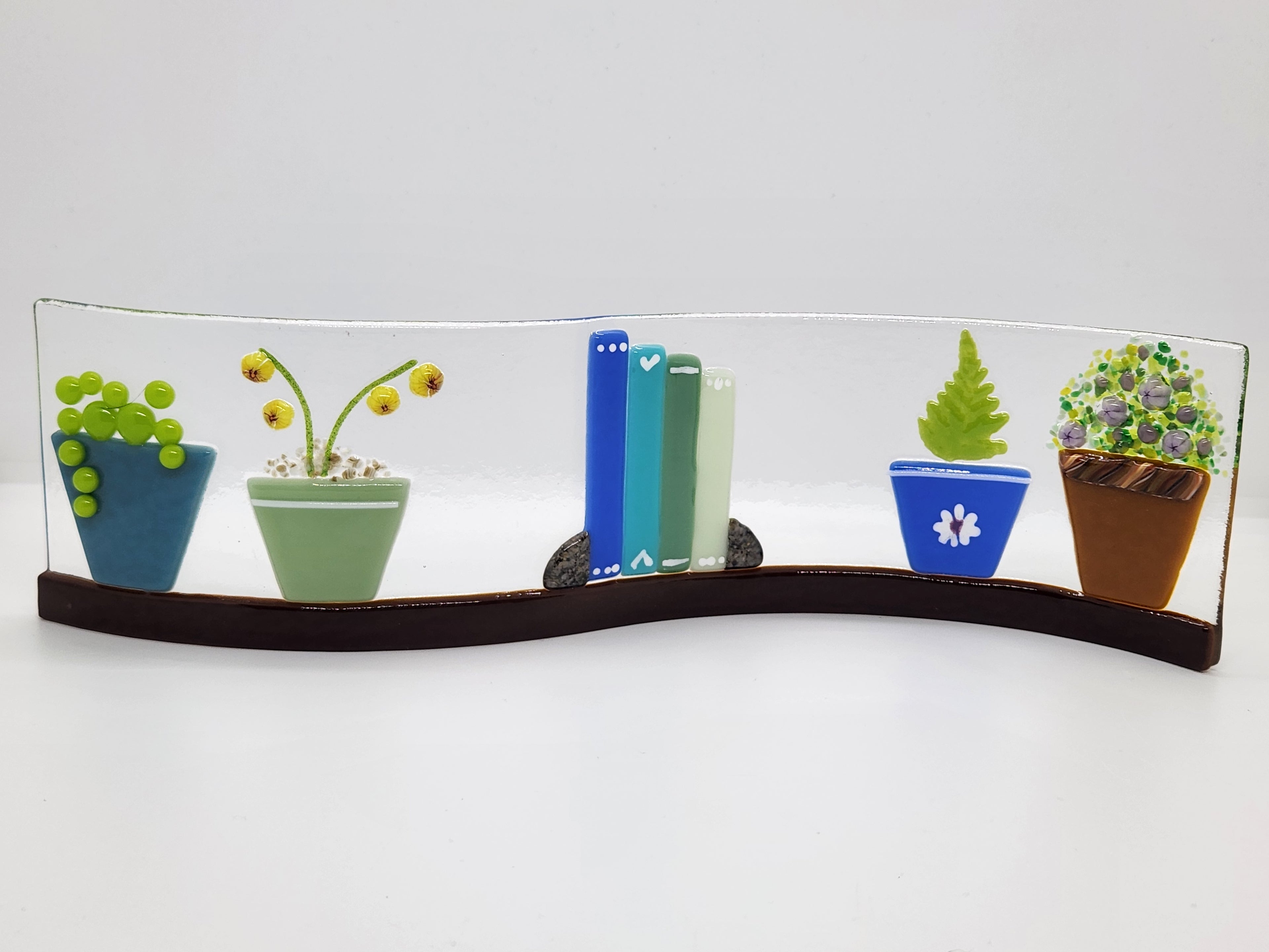 Fused glass Book Shelf Wave featuring colorful books, potted plants, and garden-inspired details on a gently curved glass panel for home decor.