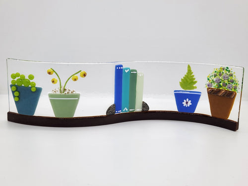 Fused glass Book Shelf Wave featuring colorful books, potted plants, and garden-inspired details on a gently curved glass panel for home decor.