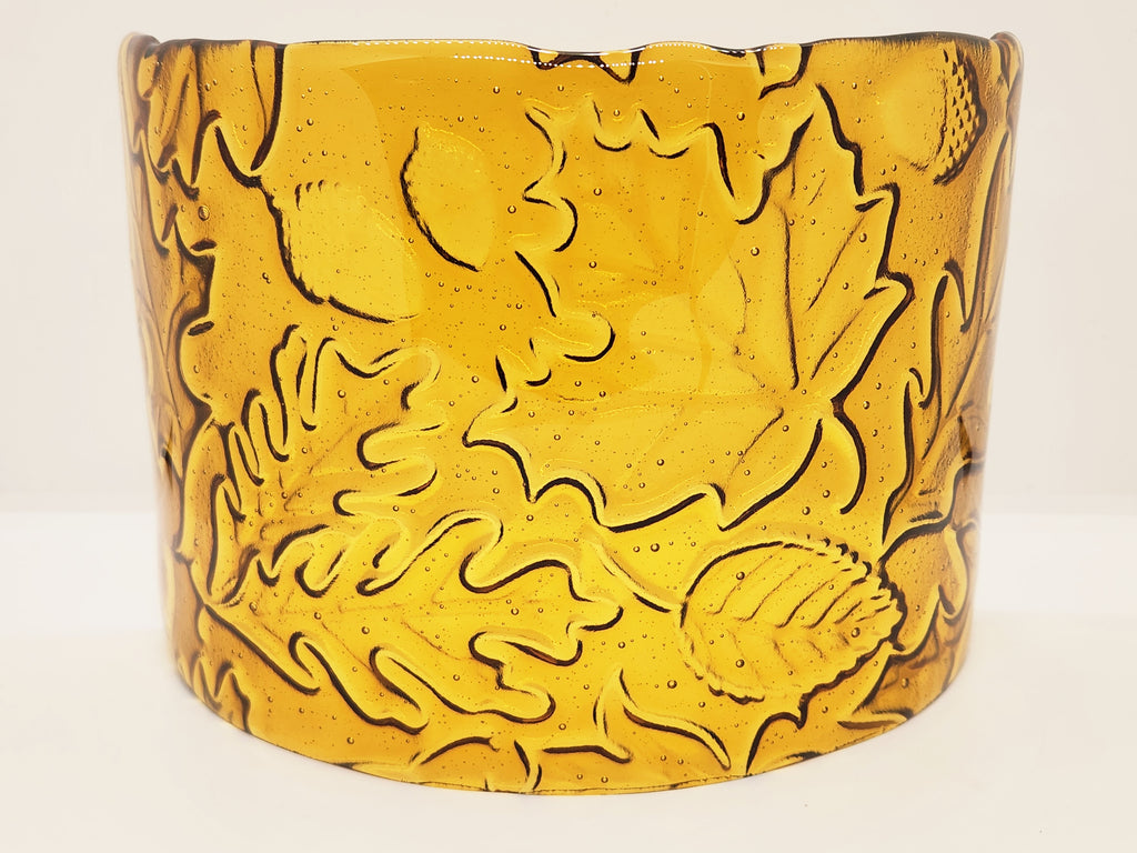 An amber glass candle holder featuring a raised relief pattern of autumn leaves and acorns, shown against a plain light background.