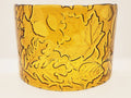 An amber glass candle holder featuring a raised relief pattern of autumn leaves and acorns, shown against a plain light background.