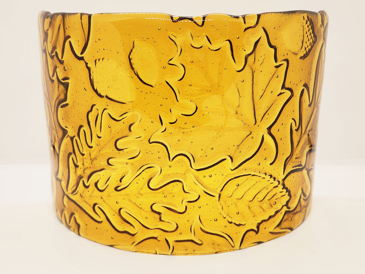 An amber glass candle holder featuring a raised relief pattern of autumn leaves and acorns, shown against a plain light background.