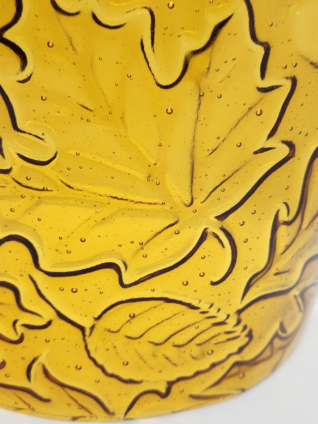 An amber glass candle holder featuring a raised relief pattern of autumn leaves and acorns, shown against a plain light background.