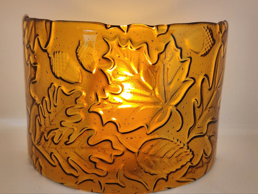 An amber glass candle holder featuring a raised relief pattern of autumn leaves and acorns, shown against a plain light background.