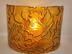 An amber glass candle holder featuring a raised relief pattern of autumn leaves and acorns, shown against a plain light background.