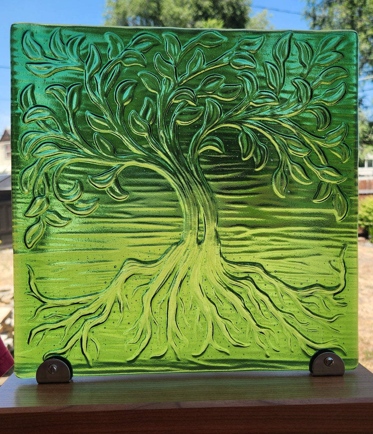 A square glass art tile featuring a raised, textured tree design in iridescent green tones, displayed on a light wood stand outdoors.