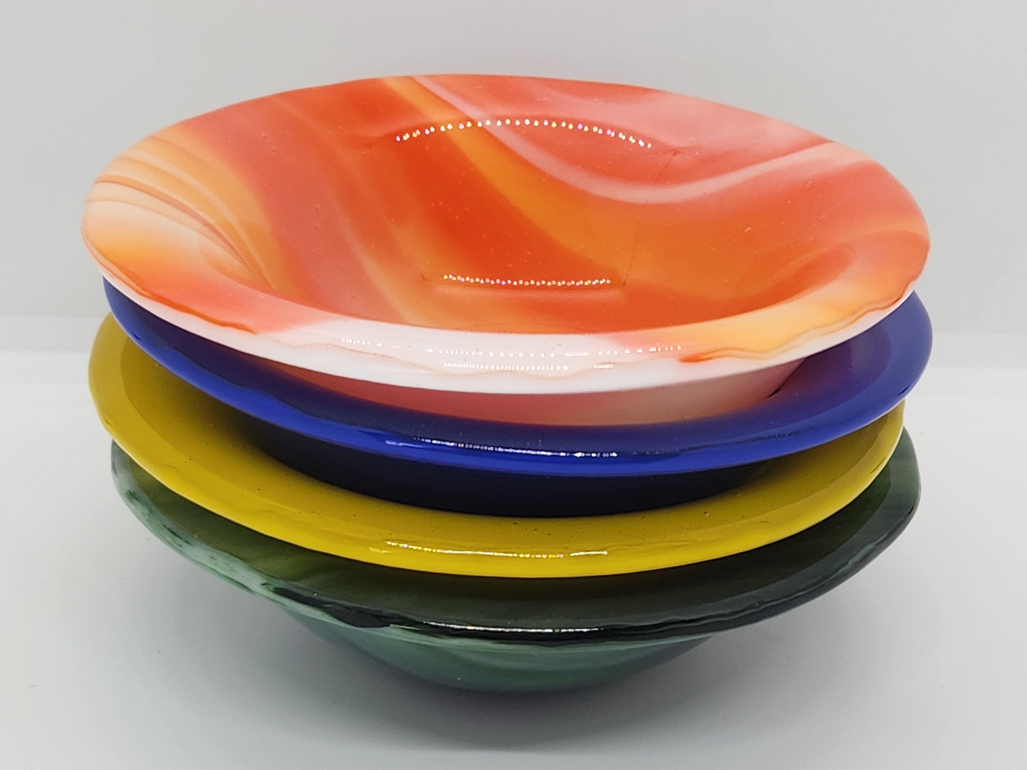 A stack of four hand-fused glass bowls in vibrant colors: orange and white marbled on top, followed by blue, yellow, and dark green.