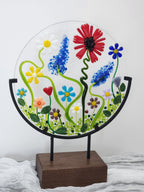 A circular clear glass wall art piece featuring colorful fused flowers and green vines, mounted on a black metal frame attached to a rectangular wooden stand.