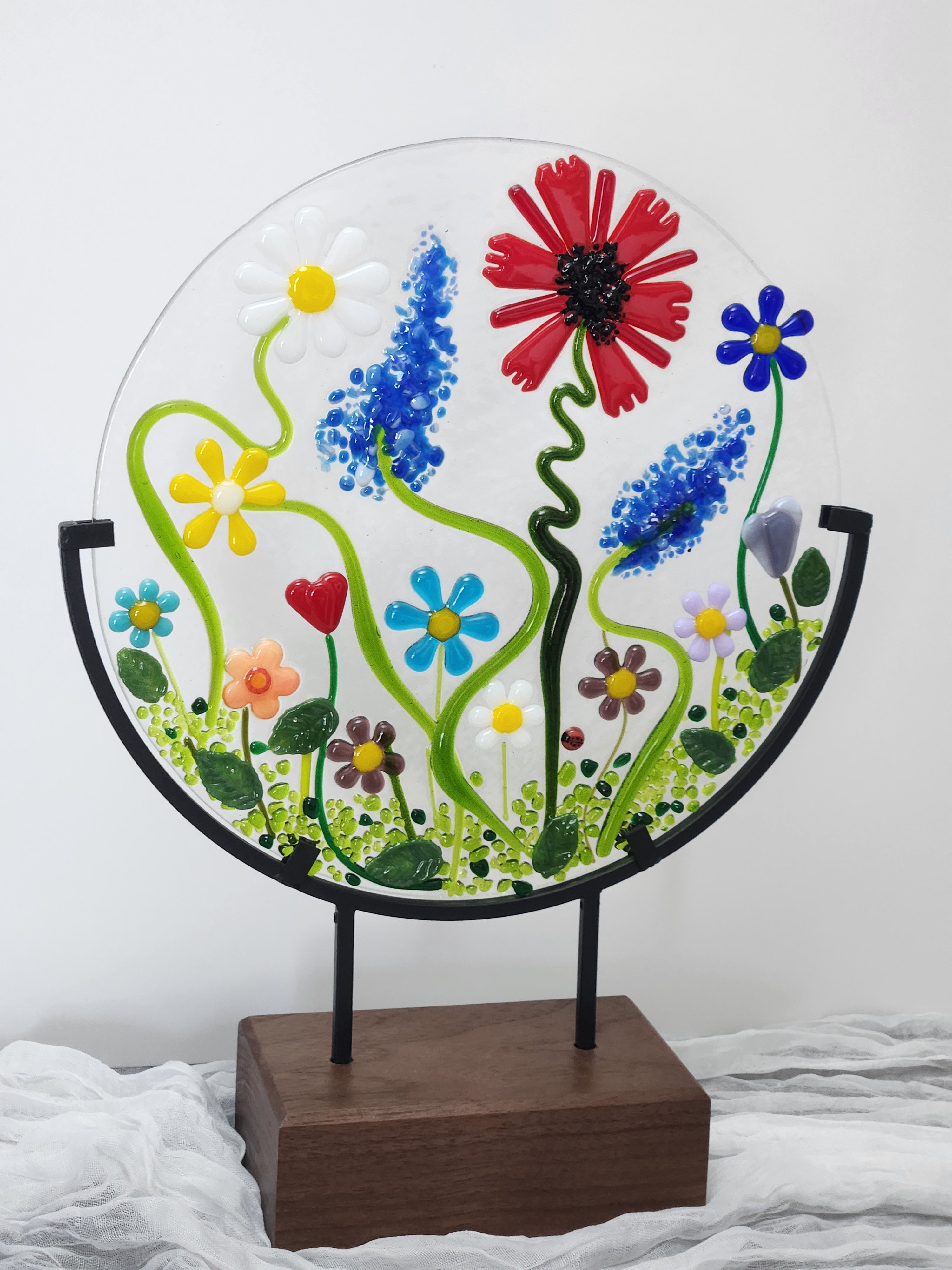 A circular clear glass wall art piece featuring colorful fused flowers and green vines, mounted on a black metal frame attached to a rectangular wooden stand.