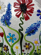 A circular clear glass wall art piece featuring colorful fused flowers and green vines.
