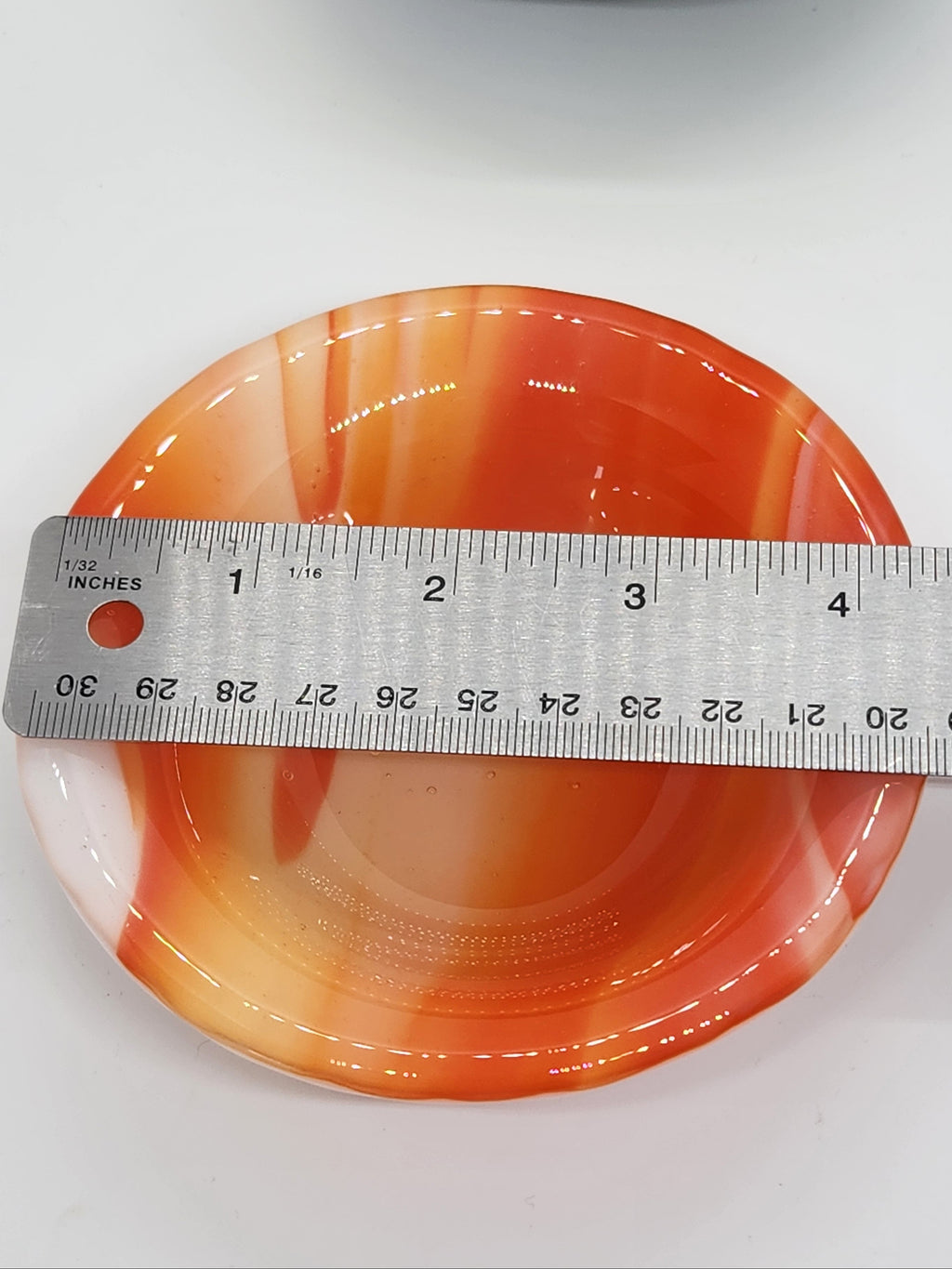 An orange and white marbled glass trinket dish shown with a metal ruler to indicate its size.