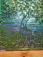 A square glass art tile featuring a raised, textured tree design in iridescent green, blue, and purple tones, displayed on a light wood stand.