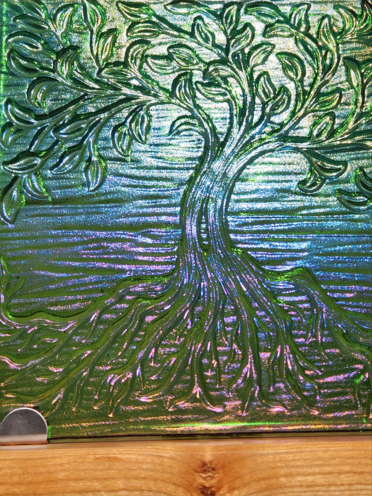 A square glass art tile featuring a raised, textured tree design in iridescent green, blue, and purple tones, displayed on a light wood stand.
