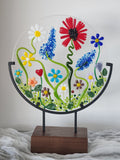 A circular clear glass wall art piece featuring colorful fused flowers and green vines, mounted on a black metal frame attached to a rectangular wooden stand.