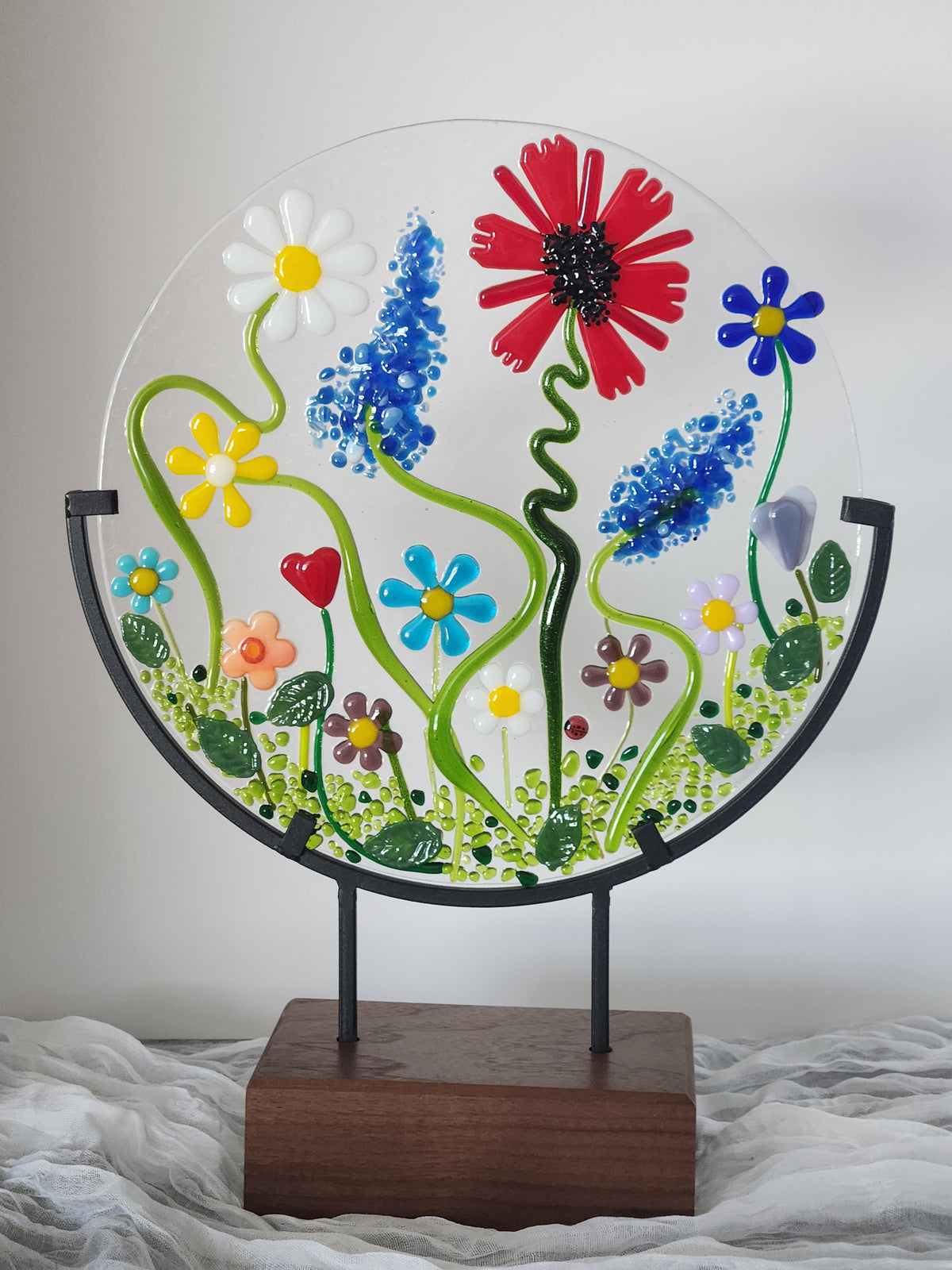A circular clear glass wall art piece featuring colorful fused flowers and green vines, mounted on a black metal frame attached to a rectangular wooden stand.